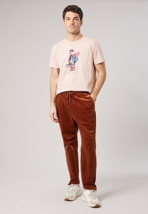 Man staat met hand in zak, draagt een beige t-shirt met teddybeerprint, roestkleurige ribfluwelen broek en witte sneakers.