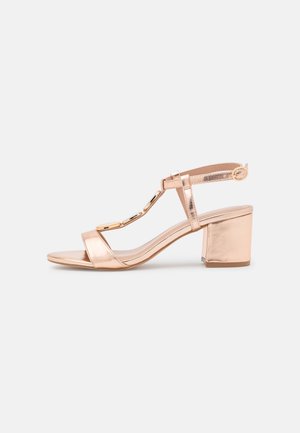 Rosen-guld hæl sandal med blokhæle, brede stropper og en dekorativ kædeaccent på forsiden. Glat metallisk finish.