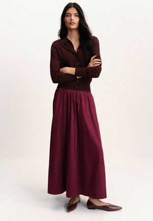 Frau mit langen dunklen Haaren, die eine burgunderfarbene Strickjacke, einen passenden langen Rock und braune spitze Ballerinas trägt, steht mit verschränkten Armen.