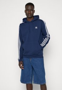Marinblå huvtröja med vita sidorand, features en framficka och en liten broderad logo. Kombination med blå denimshorts.
