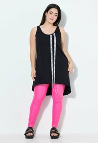 Top nero senza maniche con una striscia bianca con il marchio al centro, abbinato a leggings rosa lucidi e sandali neri con zeppa.