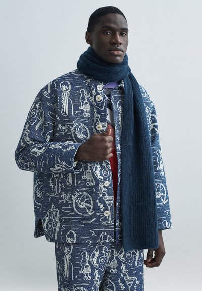Veste bleue à motifs avec des griffonnages blancs, dotée de gros boutons, accompagnée d'une écharpe bleu foncé. Le tissu présente une apparence texturée.