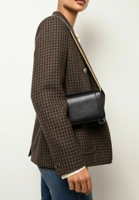 Fekete bőr keresztbody táska téglalap alakú, lánc pánttal és elegáns dizájnnal, egy barna kutyafogmintás blézer felett viselve.