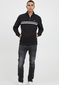 Black zip-up sweatshirt with grey chest stripes, featuring a stand collar, ribbed cuffs, and a soft fabric texture. Paired with dark jeans and white shoes.