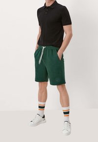 Shorts en coton verts avec une taille élastique et un cordon blanc, assortis à un polo noir et des baskets blanches. Chaussettes blanches à rayures.