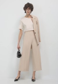 Pantalon beige sur mesure, top en soie crème, et blazer assorti. Talons en vernis noir et sac à main. Textures lisses, design structuré et lignes épurées.