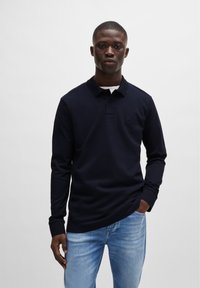 Marineblauwe poloshirt met lange mouwen en een kraag, voorzien van een subtiele logo patch, gecombineerd met lichtblauwe jeans. Soepele katoenstructuur.