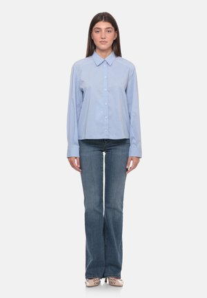 Donna che indossa una camicia azzurro chiaro con bottoni e piccoli puntini, jeans blu a zampa e scarpe beige con tacco, in piedi davanti a uno sfondo semplice.