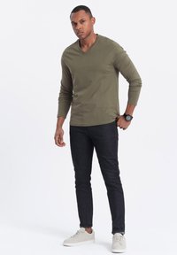 Olivgrünes langärmliges V-Ausschnitt-Shirt, getragen mit dunkelblauen Jeans. Das Modell trägt hellfarbige Sneakers und eine schwarze Uhr. Schlichtes, tailliertes Design.