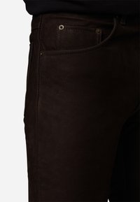 Donkerbruin jeans met een gladde textuur, traditionele zakken, zichtbare stiksels en metalen hardwareaccenten op de taille.