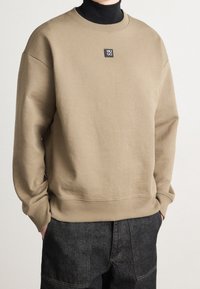 Uomo che indossa un felpa beige Hugo sopra un dolcevita nero, con le mani nelle tasche di pantaloni in denim scuro, in piedi contro uno sfondo neutro.