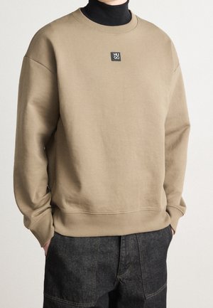 Uomo che indossa un felpa beige Hugo sopra un dolcevita nero, con le mani nelle tasche di pantaloni in denim scuro, in piedi contro uno sfondo neutro.