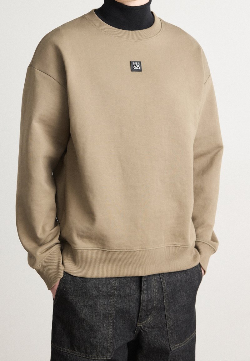 Uomo che indossa un felpa beige Hugo sopra un dolcevita nero, con le mani nelle tasche di pantaloni in denim scuro, in piedi contro uno sfondo neutro.