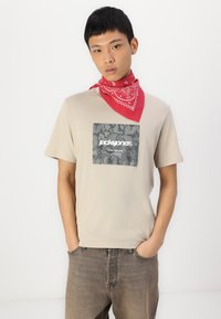 Camiseta beige de manga corta con un gráfico de camuflaje gris en forma de cuadrado y texto blanco. Llevada con un pañuelo rojo alrededor del cuello.