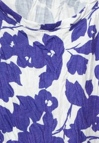 Bílá látka s velkým fialovým květinovým vzorem a jemnou vertikální texturou, ukazující zakřivený okraj výstřihu.