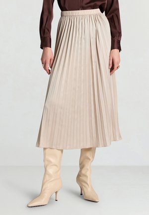 Femme portant une jupe midi plissée crème associée à des bottes à talons hautes beiges et un chemisier manches longues marron foncé.