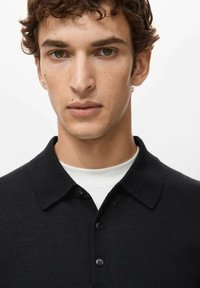 Giovane uomo con capelli castani ricci e occhi nocciola, che indossa una camicia nera con colletto sopra una maglietta bianca girocollo, su uno sfondo semplice.