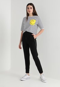 T-shirt às riscas em branco e preto com um detalhe de bolso de smiley amarelo, combinada com calças de treino pretas com riscas laterais e ténis brancos.
