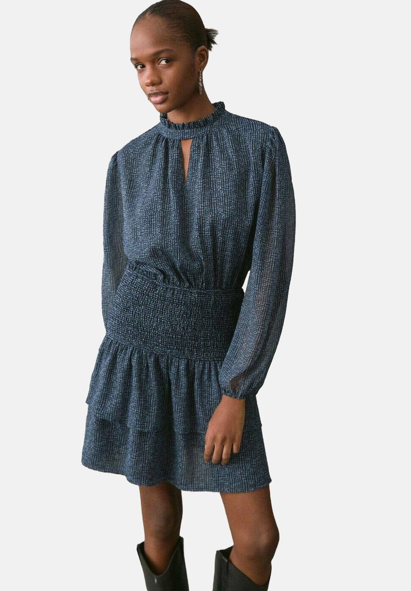 Vestido de un azul oscuro con textura, cintura fruncida, mangas largas y un dobladillo con volantes. Detalle de escote con abertura. Par de botas negras.