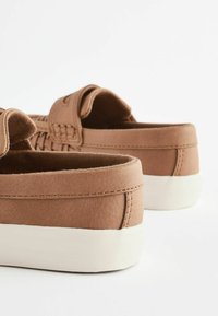 Brązowe zamszowe buty wsuwane na białej gumowej podeszwie, z teksturowaną cholewką, miękką wyściółką i kontrastującymi detalami w szwie.