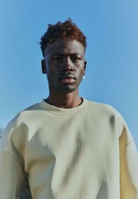 Sweat-shirt beige ample avec un col rond. Le tissu semble doux, avec des manches courtes et une silhouette décontractée.