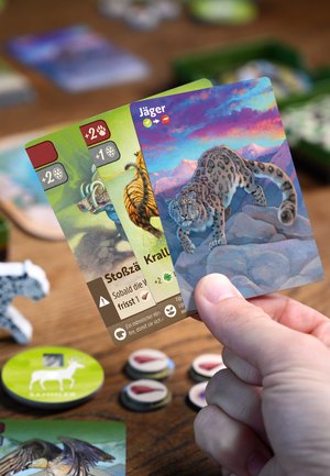 Hand holding three animal-themed game cards showing a snow leopard, tiger, and musk ox with game tokens on a wooden table.