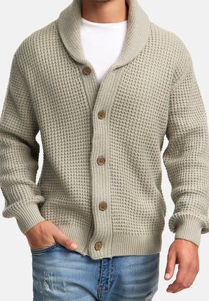 Mann trägt eine hellgraue, strukturierte Strickjacke mit Holzknöpfen über einem weißen Hemd und blau zerstörten Jeans.