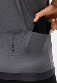 Χέρι που βάζει ένα μαύρο smartphone στην πίσω τσέπη μιας γκρι ποδηλατικής φανέλας Castelli με υφασμάτινη υφή.