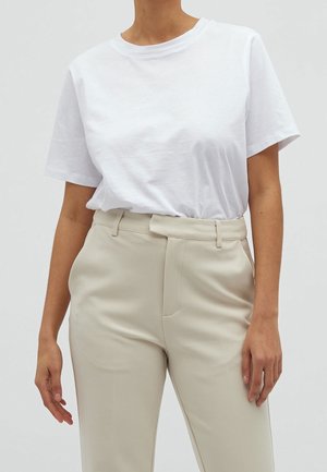 Vrouw draagt een eenvoudige witte T-shirt met korte mouwen, ingestopt in beige, hoog getailleerde, op maat gemaakte broek tegen een neutrale achtergrond.