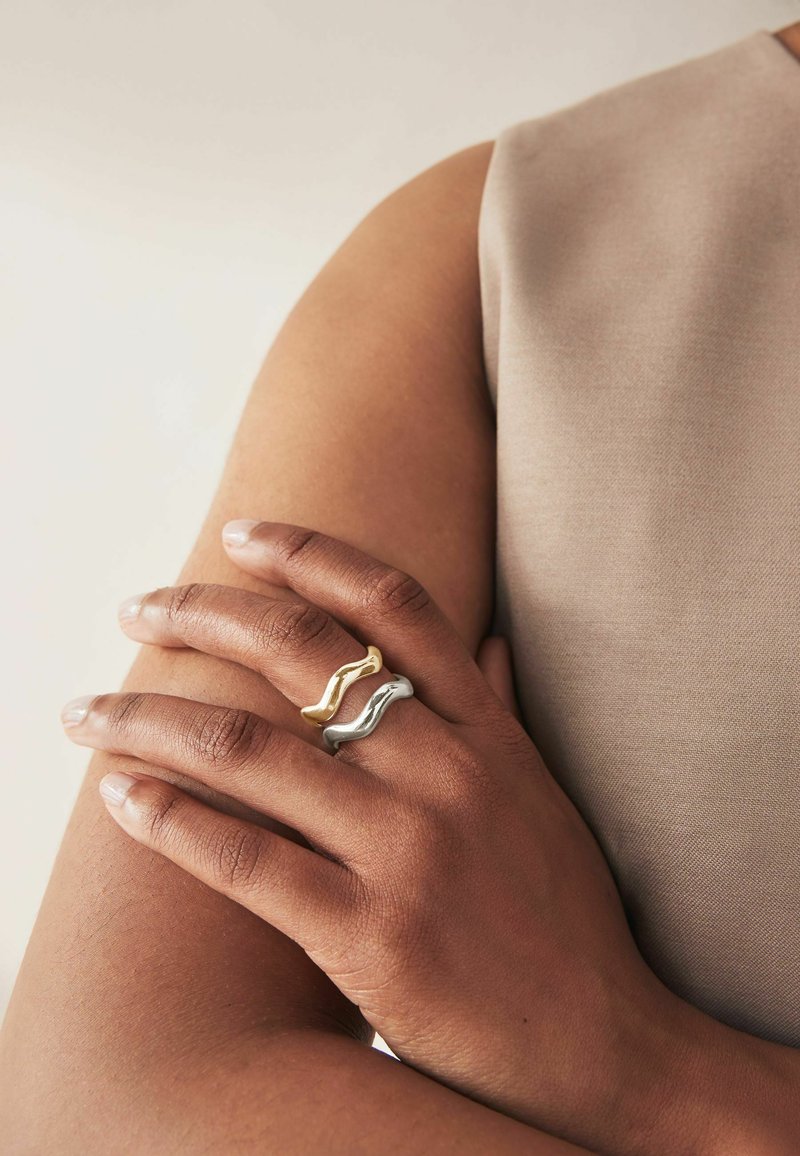 Zwei wellenförmige Ringe, ein goldener und ein silberner, getragen an Fingern neben einem nackten Arm. Die Ringe haben eine glatte, polierte Oberfläche und eine einzigartige Form.