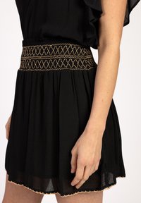 Zwarte jurk met elastische tailleband en ingewikkelde gouden borduursels. Vloeiende stof, korte lengte en delicate gouden rand bij de zoom.