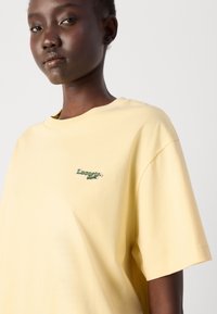 T-shirt en coton jaune avec une coupe ample, décoré d'un petit logo Lacoste brodé en vert sur la poitrine. Texture douce et design simple.