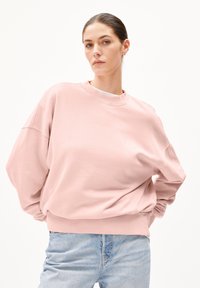 Rosa oversized sweatshirt med rund hals och sänkta axlar. Mjuk textur med diskret branding nära kragen. Matcher med ljusblå jeans.