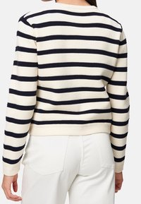 Pull crème avec des rayures horizontales bleu marine, texture côtelée, encolure ronde et ourlet ajusté. Porté avec un pantalon blanc.