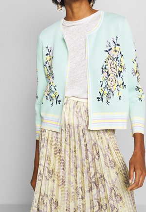 Femme portant un cardigan bleu clair brodé de fleurs sur un t-shirt blanc, associé à une jupe plissée à imprimé serpent.