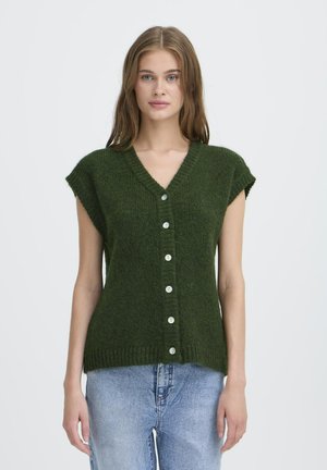 Woman wearing a green knit sleeveless buttoned cardigan and light blue jeans, standing against a plain light background.