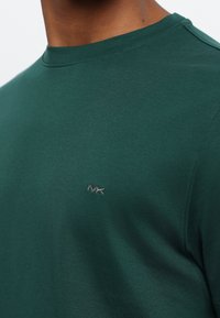 Žalias medvilninis marškinėliai su apykakle, kuriame yra mažas pilkas siuvinėtas logotipas kairėje krūtinės pusėje ir matomi siuvimo detalės.