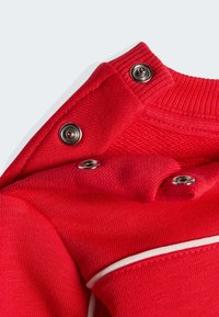 Crveni odjevni predmet od tkanine s teksturiranom površinom, rebrastim okovratnikom i srebrnim kopčama na pritisak. Naglašeni detalji uključuju bijelo obrubljene šavove.