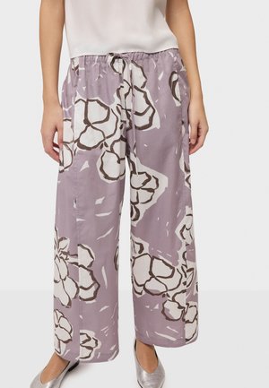 Žena má na sebe široké levanduľové nohavice s veľkým bielym a hnedým kvetinovým vzorom, elastický pás so sťahovacou šnúrkou a strieborné baleríny.