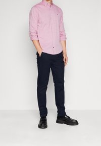 Helles rosa Hemd mit einem subtilen Muster, kombiniert mit dunkelblauen Hosen und schwarzen Mokassins. Das Hemd hat einen Kragen und eine einzelne Brusttasche.