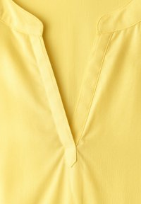 Col de chemise jaune en V avec une courte patte de boutonnage et des coutures visibles sur un tissu lisse.