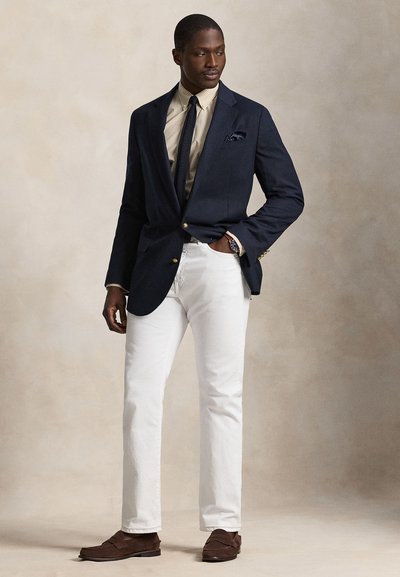 Homme portant un blazer bleu marine, une chemise beige à carreaux, une cravate sombre, un pantalon blanc et des mocassins marron, debout avec une main dans la poche devant un fond neutre.