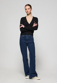 Cardigan noir avec un col en V, des manches longues et une fermeture à un bouton, associé à un jean bleu évasé taille haute et des baskets beiges.