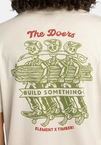 Krémové tričko s zelenou grafikou troch kostier nesúcich polená a pílou, s červeným textom "The Doers" a "BUILD SOMETHING."