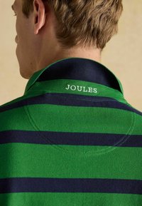 Zaļš un tumši zils svītrainas polo krekls ar apkakli, uz kura aizmugures ir izšūts teksts "JOULES" baltā krāsā. Tekstūrēts audums.