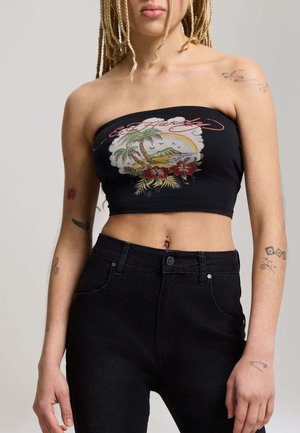 Persona con capelli intrecciati che indossa un top corto nero senza spalline con stampa tropicale e jeans neri a vita alta, mostrando diversi tatuaggi sulle braccia.