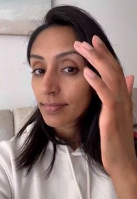 Donna con lunghi capelli neri in un hoodie bianco, che si tocca il viso con una mano. Ha un piercing al naso e un'espressione neutra.