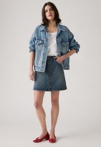 Chaqueta de mezclilla (azul claro) sobre una camiseta sin mangas blanca, combinada con una falda de mezclilla azul con botones frontales y un dobladillo sin terminar; zapatos planos rojos.
