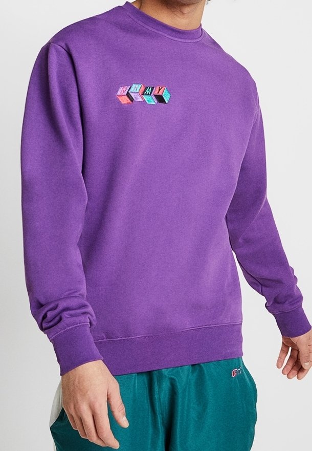 Hombre con un sudadera púrpura con letras de colores en bloque que dicen "ARMY" en el pecho y pantalones color teal.