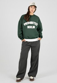 Jeune personne debout, les mains dans les poches, portant un sweat-shirt vert foncé avec l'inscription "PROHIBITED BERLIN", un pantalon gris à jambes larges, une casquette beige et des chaussures bicolores.